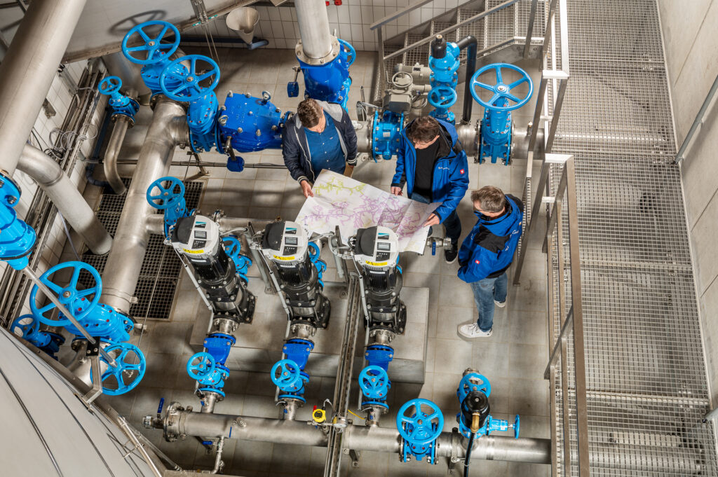 blick von oben auf drei techniker in einer wasserversorgungsanlage mit einem plan in der hand zwischen blauen rohrsystemen