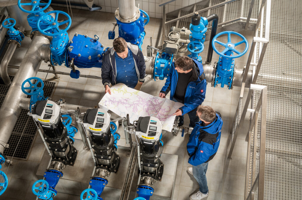 drei techniker in einer wasserversorgungsanlage mit einem plan in der hand zwischen blauen rohrsystemen